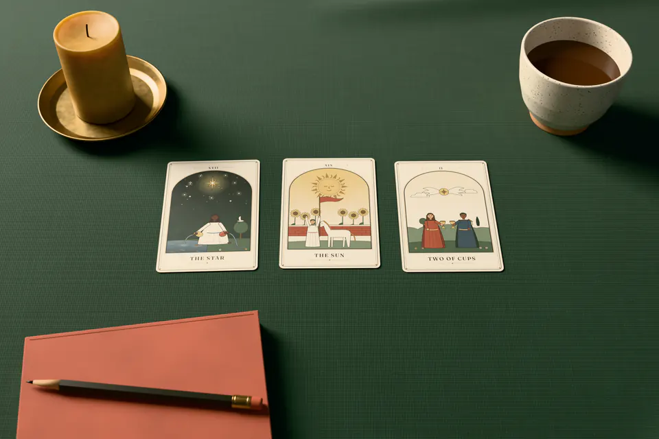 A three-card tarot spread of The Star, The Sun and Two of Cups on deep green linen, with a beeswax candle in a brass dish, a cup of tea and a notebook with a pencil, in soft window light.